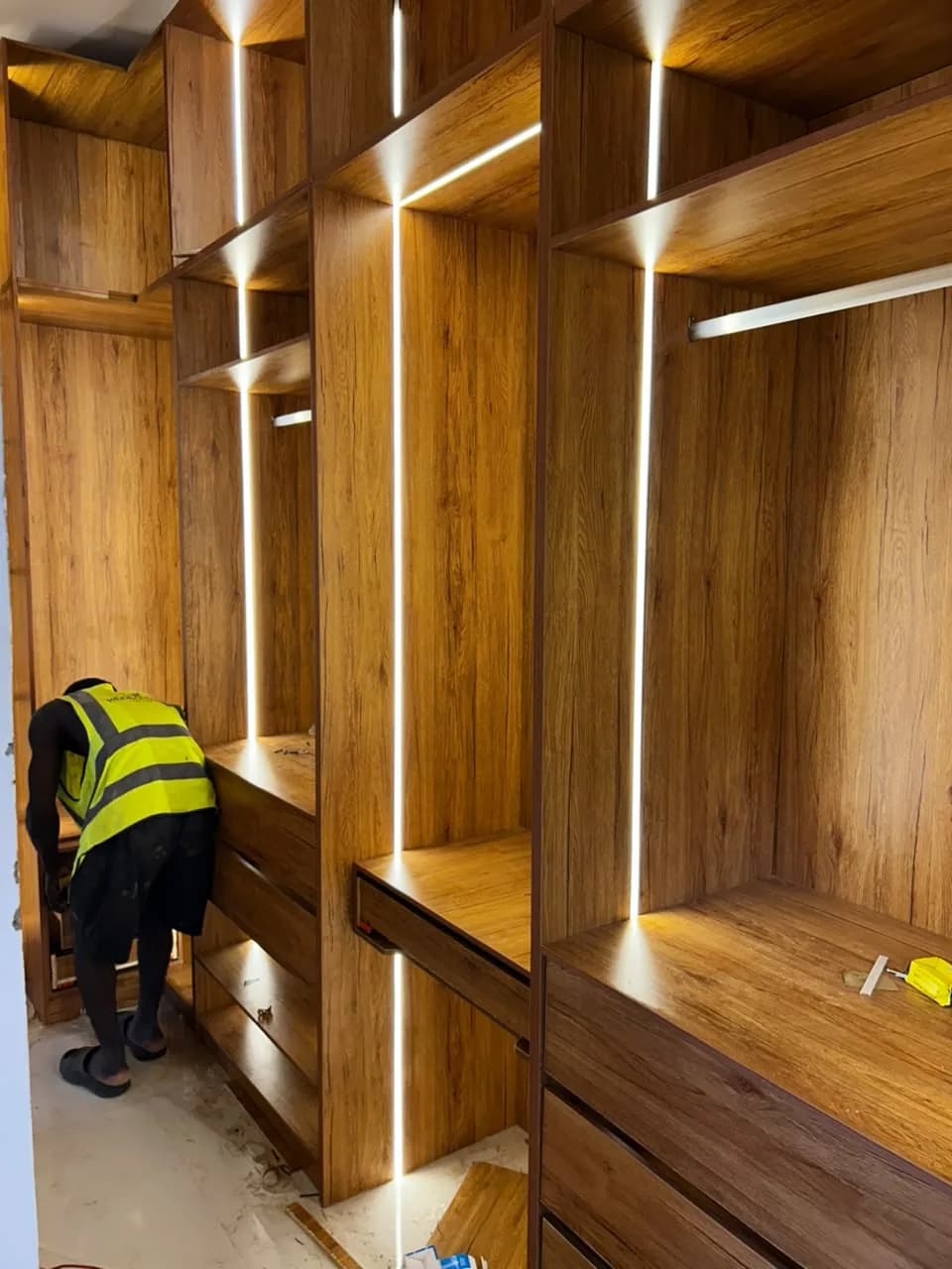 A fitter installing drawers in a walk-in timber-grain wardrobe, its shelves lit by vertical LED strips.