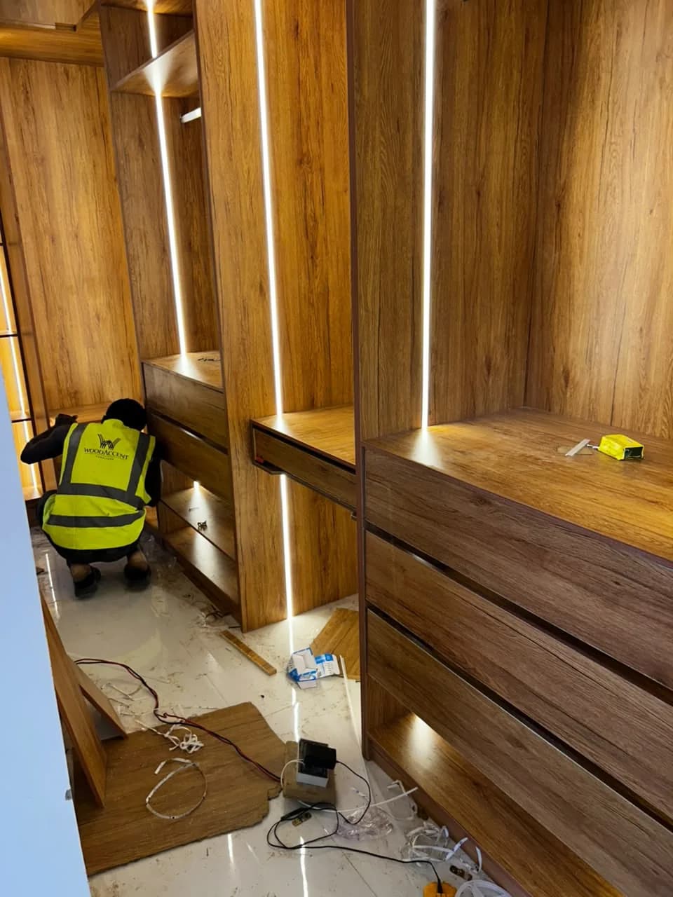 A fitter in a WoodAccent hi-vis vest crouched at the drawers of a lit walk-in timber wardrobe.