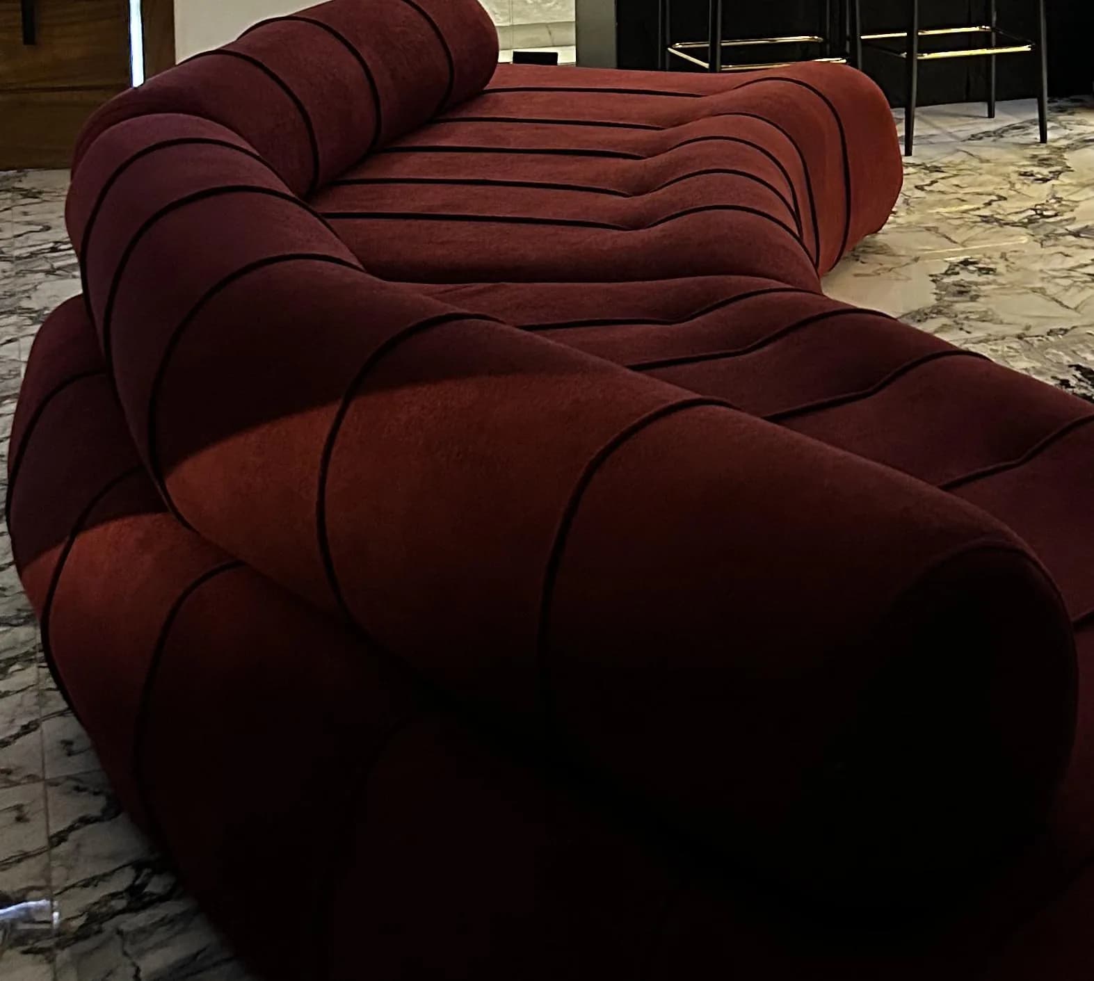 The full curve of the burgundy Helix sofa between a timber plank door, a lit bar and a brass coffee table.
