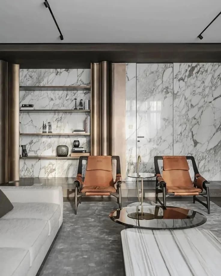 Two tan leather sling chairs on black frames between marble slab walls and brass shelving in a grey-toned room.